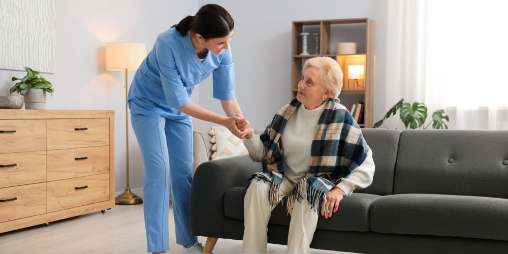 Home health care in Greenfield, IN caregiver helping an elderly woman stand up from a couch in a comfortable home setting.